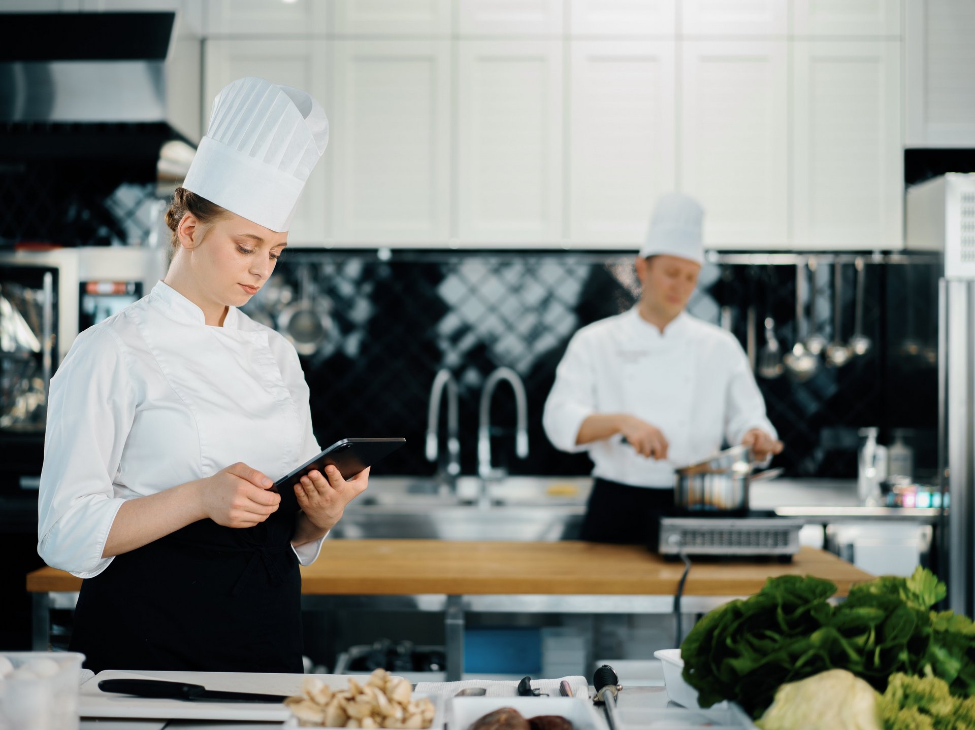 Two chefs standing in a restaurant kitchen, one is cooking and the other is using a tablet to manage digital recipes.